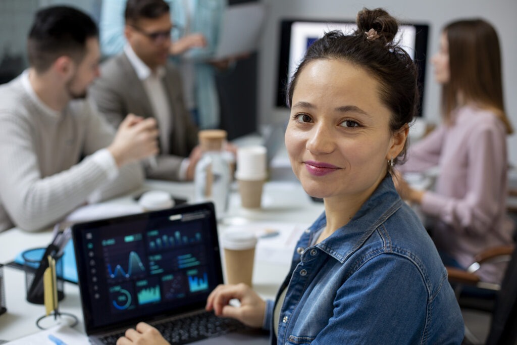 Mulher em primeiro plano com laptop aberto numa mesa de reunião.