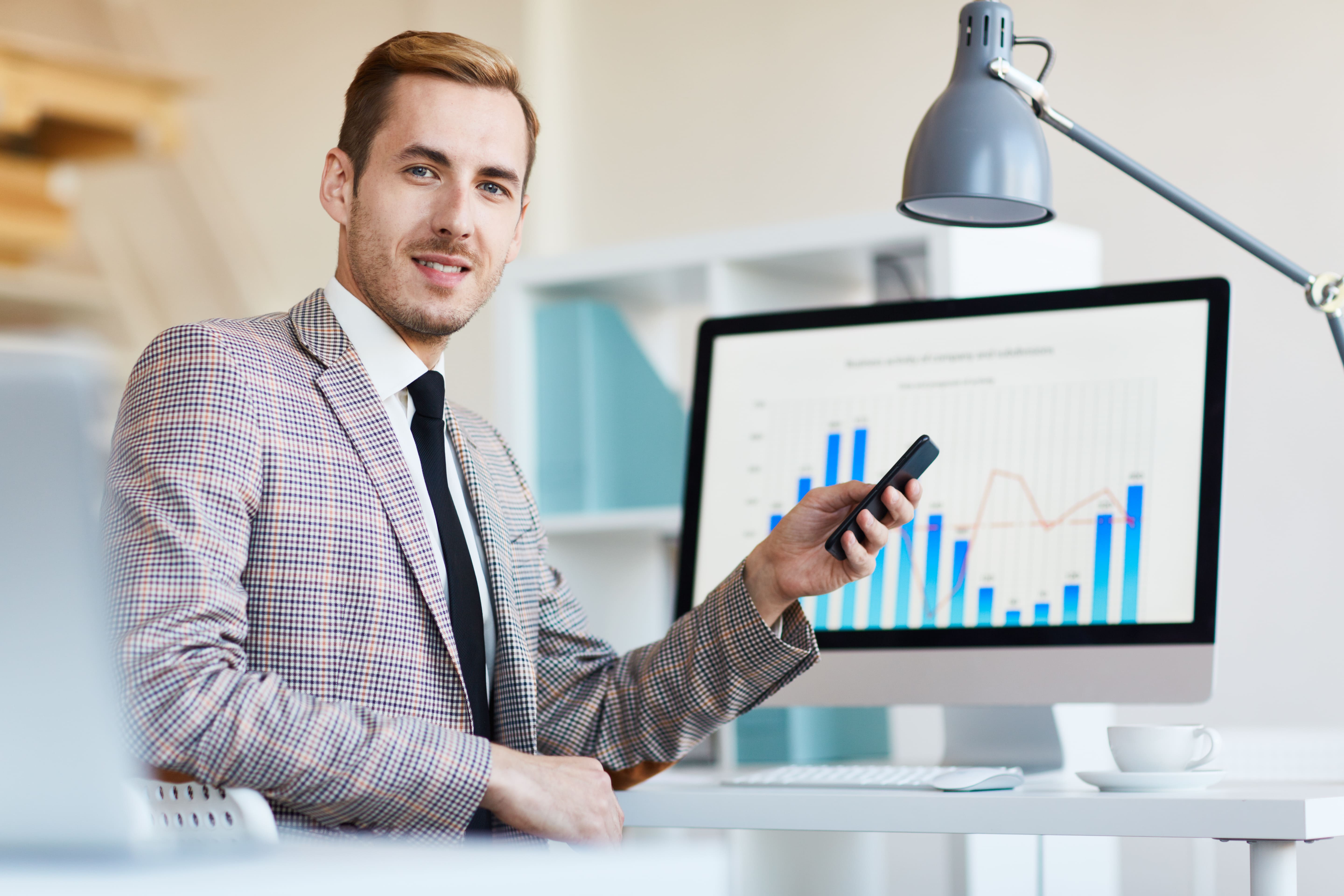 Homem jovem com celular na mão e ao fundo gráficos em uma tela.